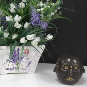 Mässingsfigur med fyra ansikten placerad bredvid en blomsterkruka med vita och lila blommor