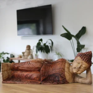 Handkarvad liggande Buddha-staty i trä på bord med växter i bakgrunden
