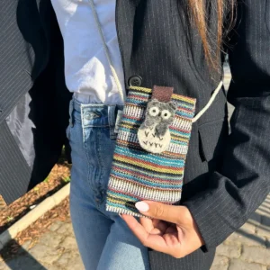 Flerfärgad mobilväska med uggla och axelrem, hålls i hand framför jeans och svart kavaj