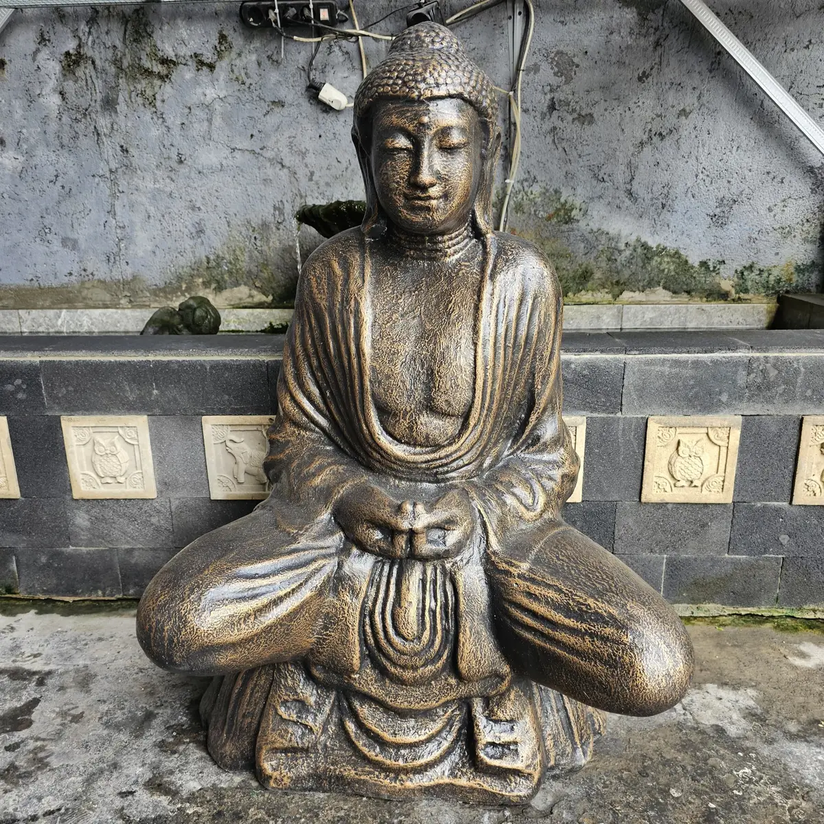 Stor antikgul buddha-staty i sittande meditationspose framför en vägg med dekorativa plattor