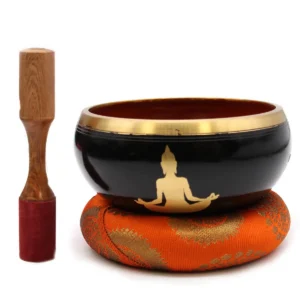 Stor svart tibetansk klangskål med guldfärgat buddha-motiv på orange kudde och träklubba