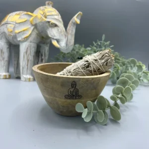 Träskål med Buddha-motiv innehållande en bunt smudge-pinnar, dekorerad med gröna blad och en vit elefantfigur i bakgrunden