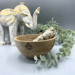 Träskål med symboler för fyra element, innehållande en smudge-pinne, med dekorativ elefantfigur och gröna blad i bakgrunden