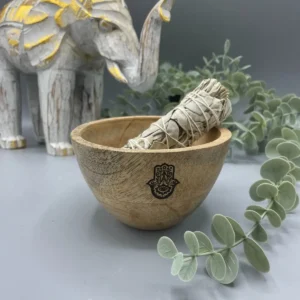 Träskål med Hamsa-symbol och smudge-pinne, omgiven av gröna blad och träfigur av elefant