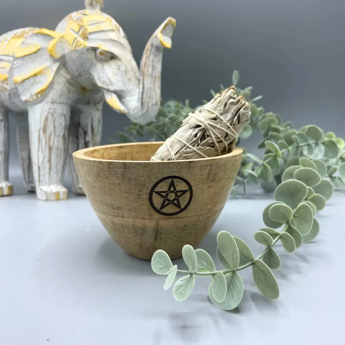 Träskål med pentagram och en bunt smudgepinnar, dekorerad med gröna blad och en vit elefantfigur i bakgrunden