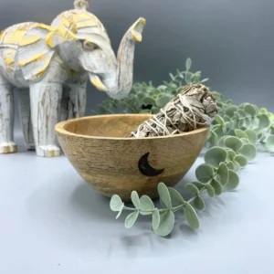Handgjord träskål med svart månmotiv och smudgepinne, omgiven av gröna blad och vit elefantfigur i bakgrunden