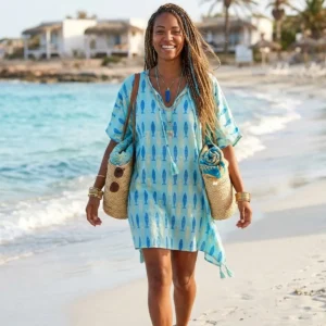 Kvinna i turkos kaftan med blå fiskmönster går på strand med hav i bakgrunden