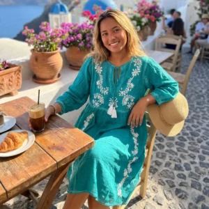Kvinna i turkos sommarklänning med vita broderier och tofsar sitter vid träbord utomhus med blommor i bakgrunden